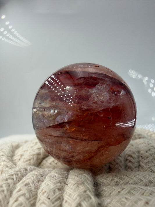 Eine hochglanzpolierte Kristallkugel mit leuchtend roten und rostfarbenen mineralischen Einschlüssen, die wie eingefrorene Flammen im klaren Quarz schweben.