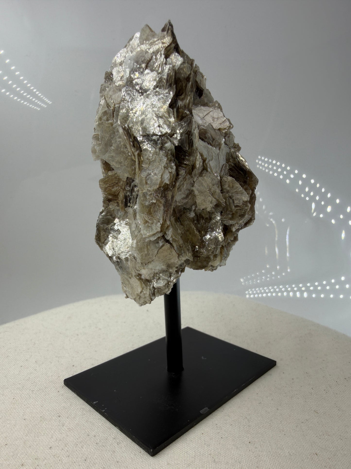 Silbrig glänzender Muskovit-Glimmer auf einem schwarzen Metallständer zur dekorativen Präsentation.