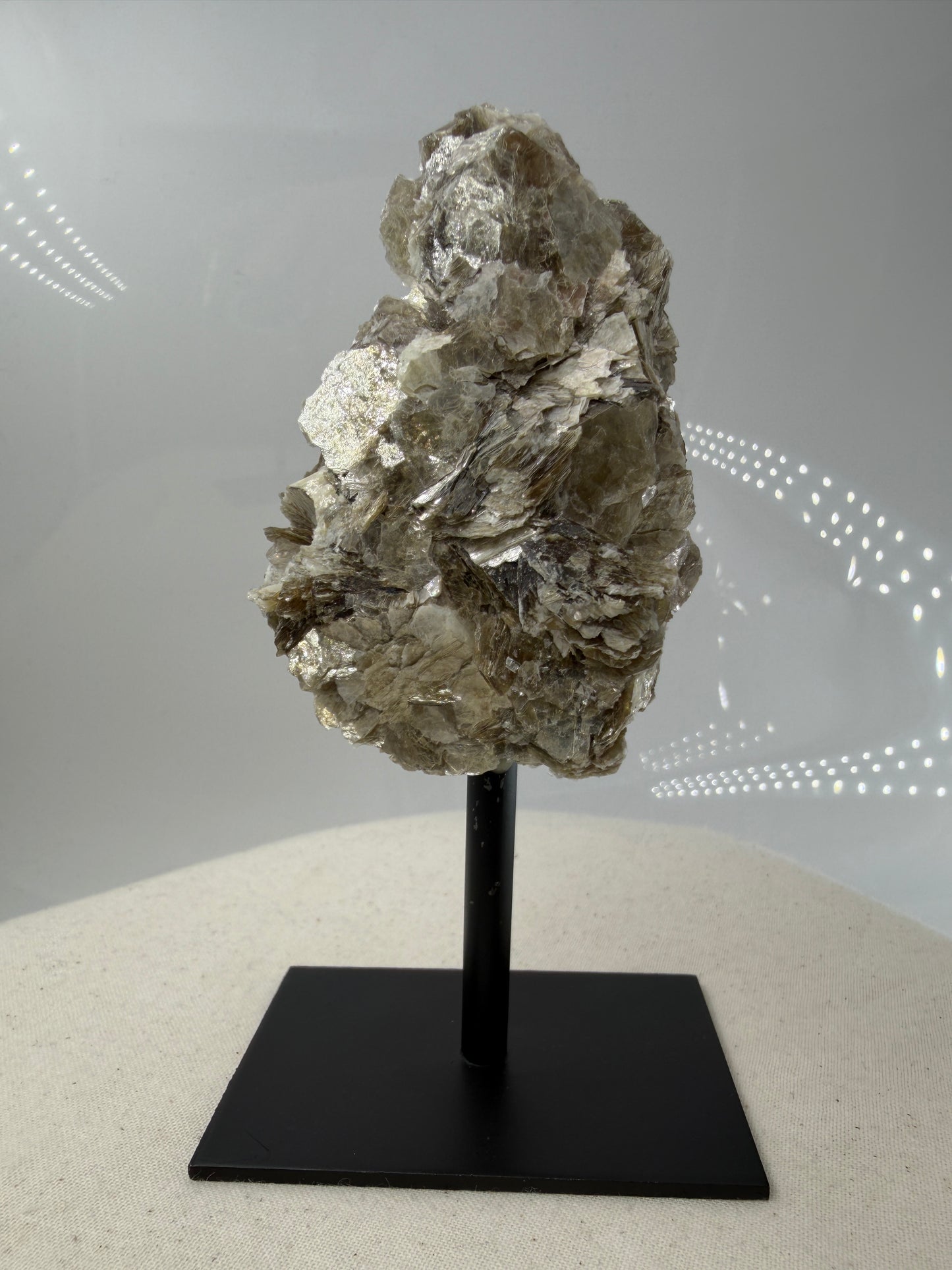 Silbrig glänzender Muskovit-Glimmer auf einem schwarzen Metallständer zur dekorativen Präsentation.