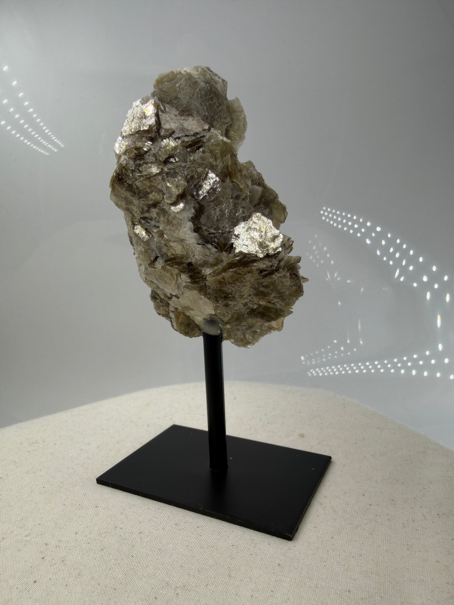 Silbrig glänzender Muskovit-Glimmer auf einem schwarzen Metallständer zur dekorativen Präsentation.