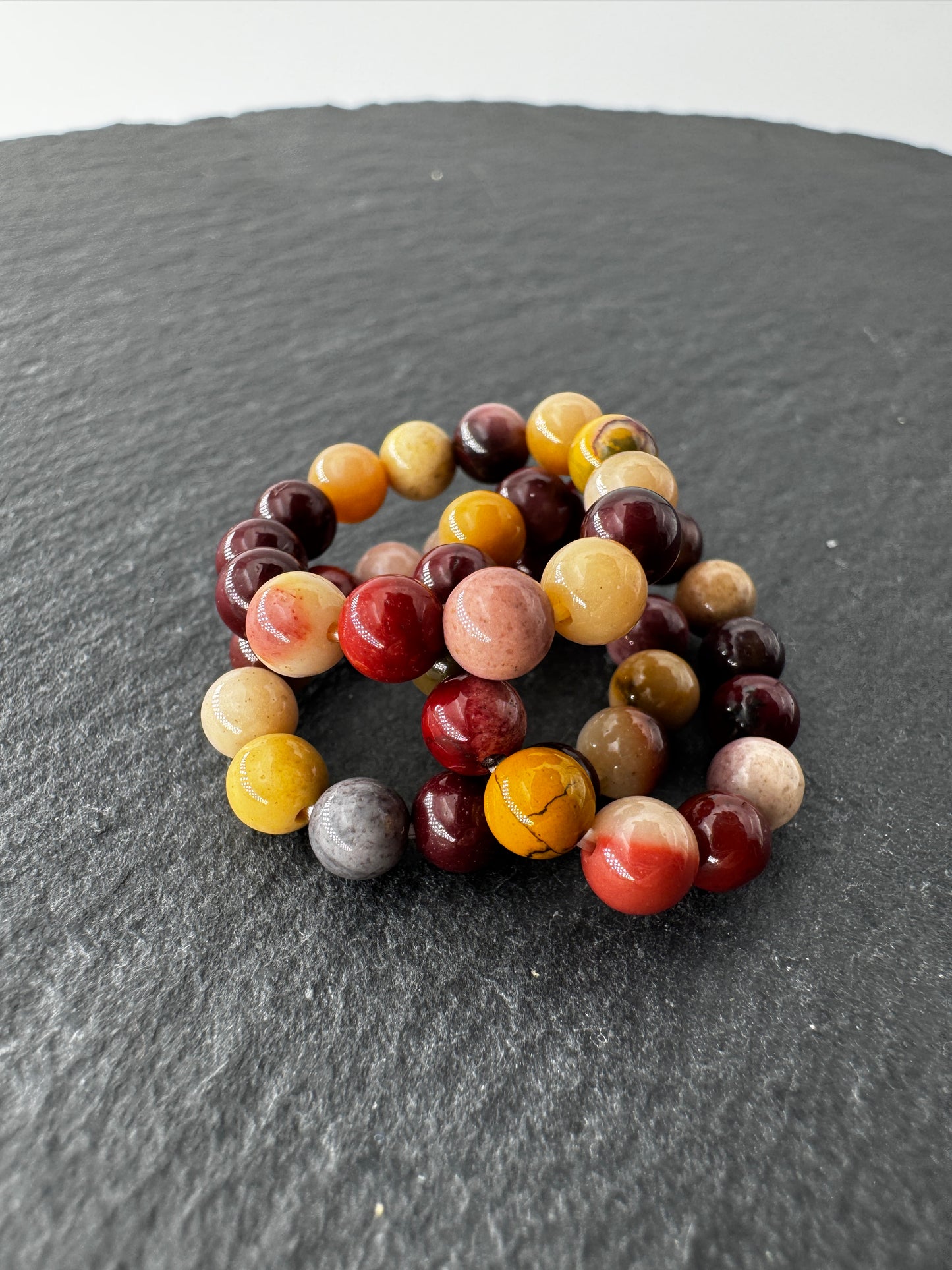 Elastischer Ring aus Mookait mit 4,6-mm-Kugeln – warme Erdtöne in Ocker, Bordeaux und Creme, Ringdurchmesser 5,5–6 cm.