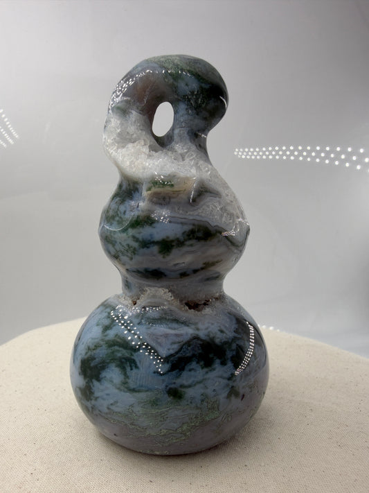 Eine polierte Skulptur in Form eines Flaschenkürbisses aus Moosachat mit tiefgrünen, moosartigen Strukturen in einer milchig-transparenten Matrix.