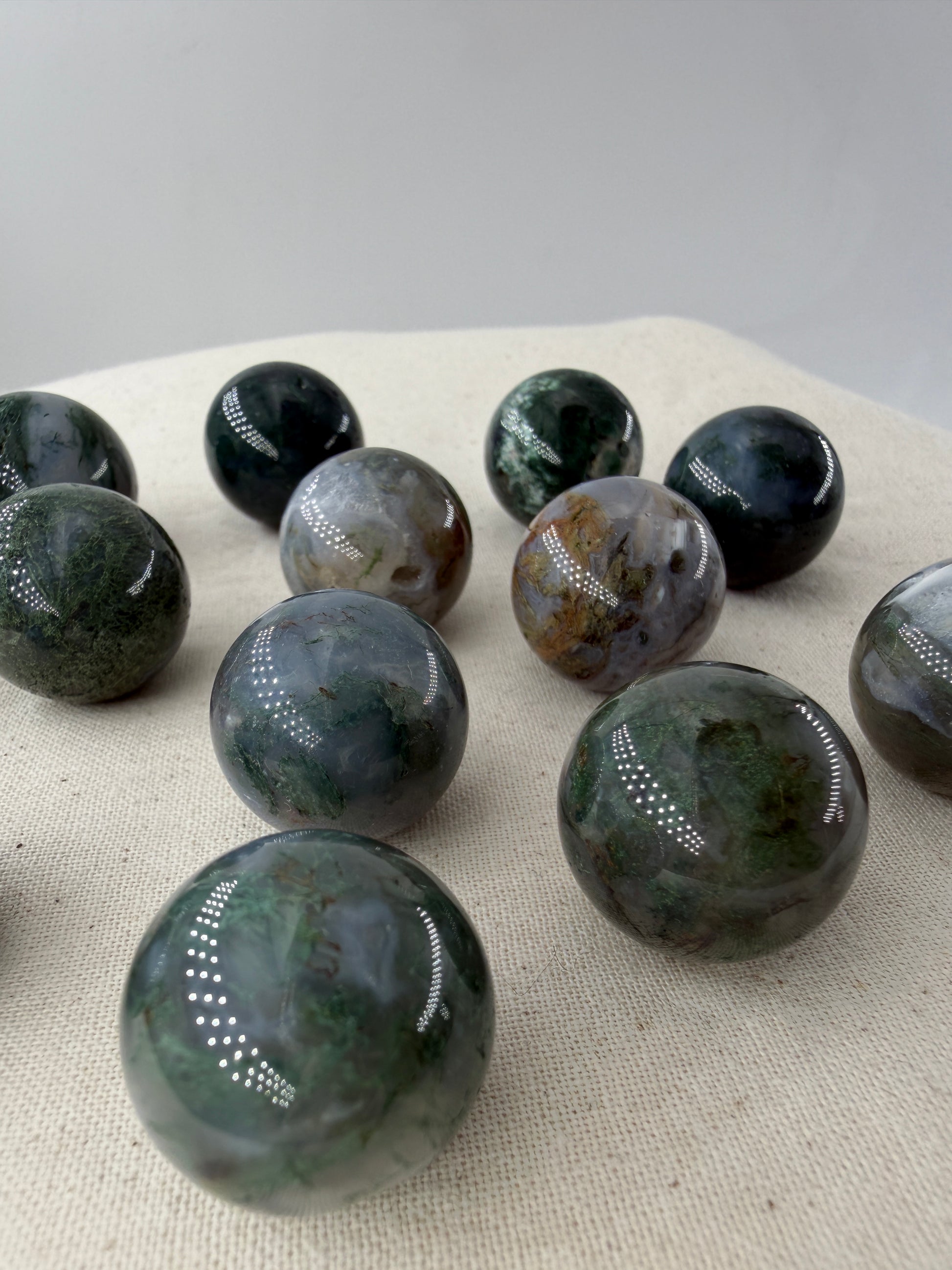 Mehrere hochglanzpolierte Minikugeln aus Moosachat mit tiefgrünen, moosartigen Strukturen in einer milchig-weißen Matrix auf einer neutralen Unterlage.