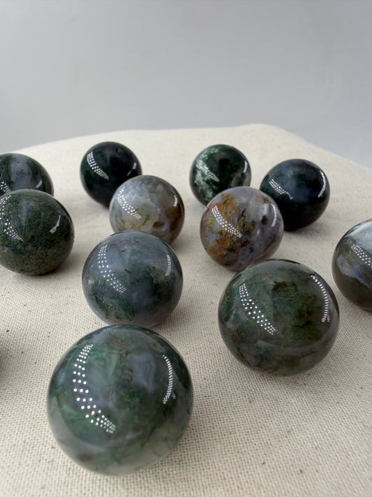 Mehrere hochglanzpolierte Minikugeln aus Moosachat mit tiefgrünen, moosartigen Strukturen in einer milchig-weißen Matrix auf einer neutralen Unterlage.