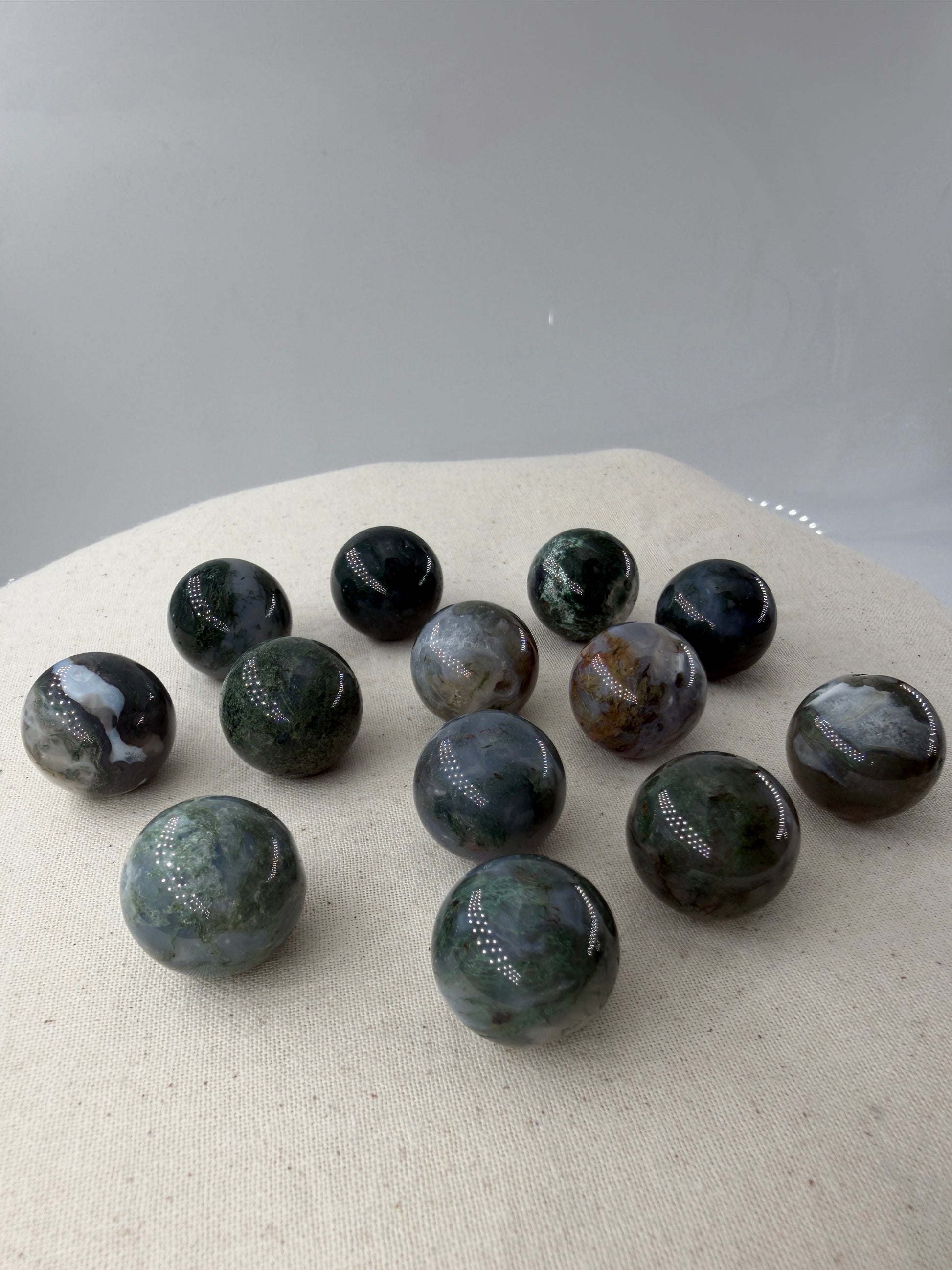 Mehrere hochglanzpolierte Minikugeln aus Moosachat mit tiefgrünen, moosartigen Strukturen in einer milchig-weißen Matrix auf einer neutralen Unterlage.