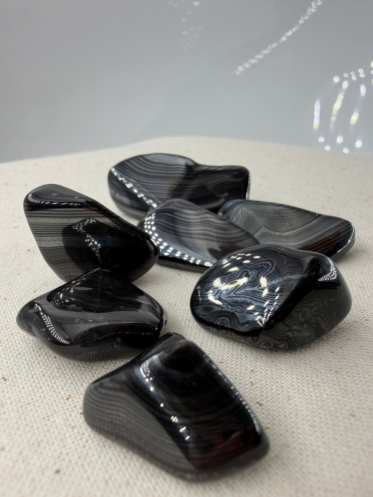 Tiefschwarze, metallisch glänzende Psilomelan-Trommelsteine mit silbrig schimmernden, fließenden Linienstrukturen auf hellem Untergrund.