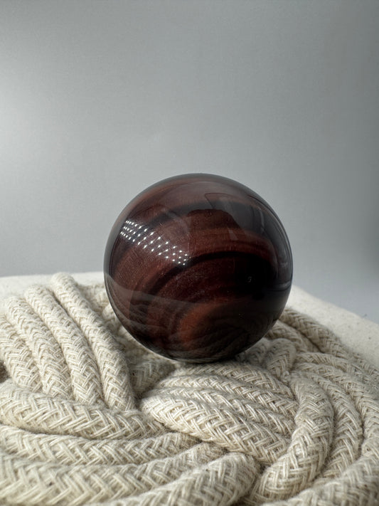 Eine perfekt polierte Kugel aus rotem Tigerauge mit einem lebendigen, rötlich-goldenen Lichtschimmer auf der dunklen Oberfläche.